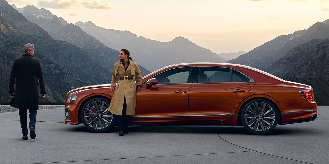 Bentley Athens Bentley Flying Spur Speed parked in Orange Flame coloured exterior parked, with mountainous background and two people in view.
